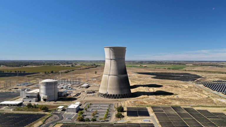Aerial view of a cooling tower surrounded by solar panels in a rural landscape.