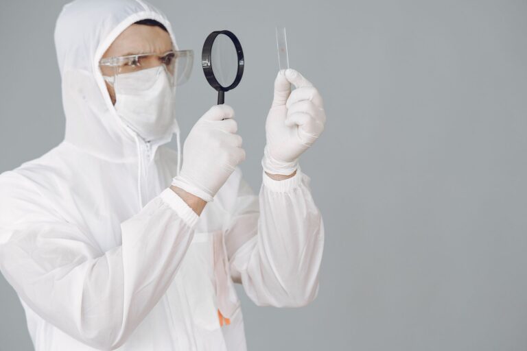 A scientist in protective gear examines a sample slide with a magnifying glass in a laboratory setting.