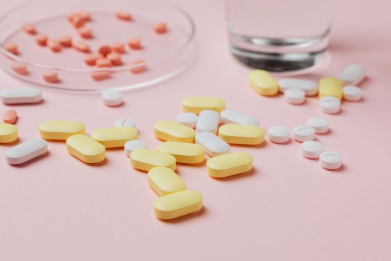 A close-up of various pills and tablets on a pink background with a glass of water.