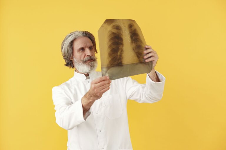 Senior male doctor holding and examining a chest x-ray, posing against a bright yellow studio background.